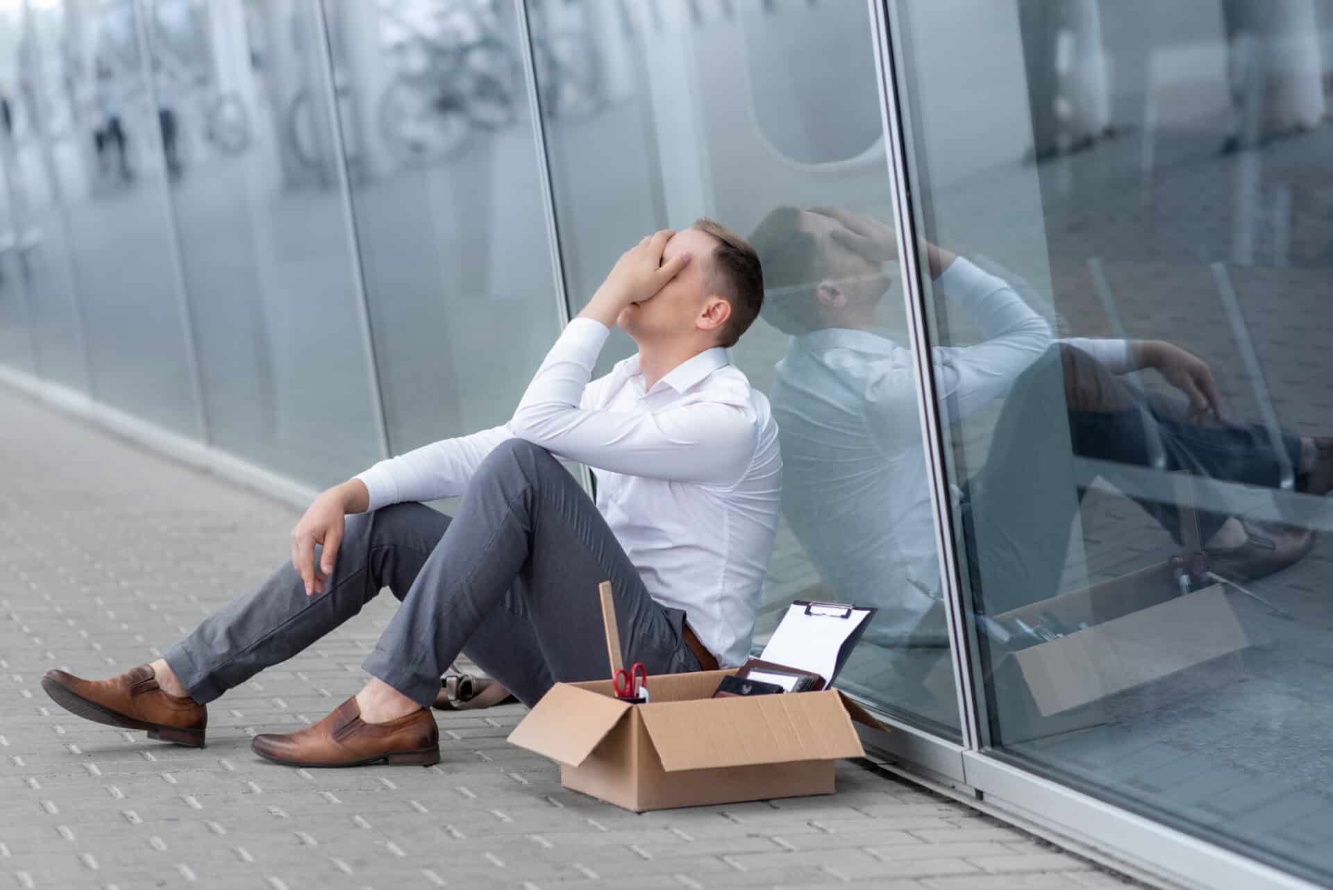 a young fired office worker is sitting under his office. next to him is his stationery.