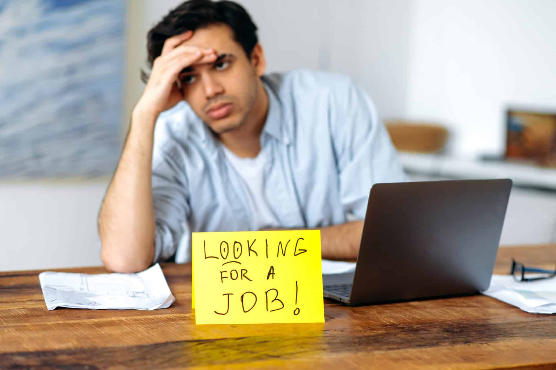Defocused desperate upset guy, student, unemployed, tired of looking for a job, sitting at his desk, looking sadly to the side, there is a sign on the table with the inscription looking for a job