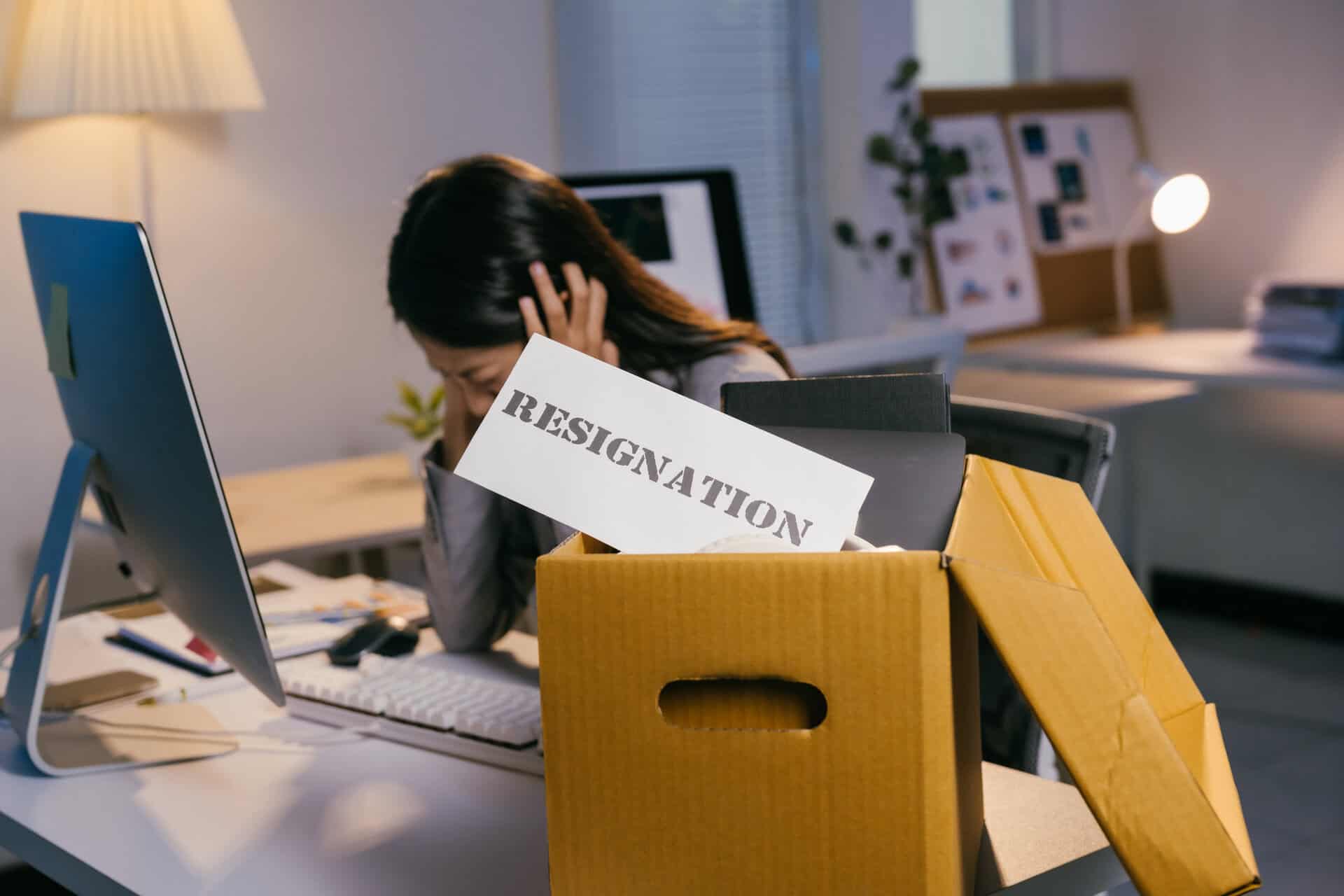 stressed businesswoman holding her head in hands while sitting at office desk with cardboard box containing her belongings and resignation letter, quitting job concept