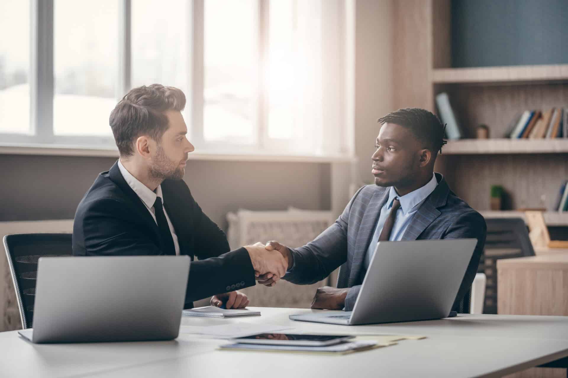 two confident businessmen in formalwear shaking hands while sitting at the desk in modern office