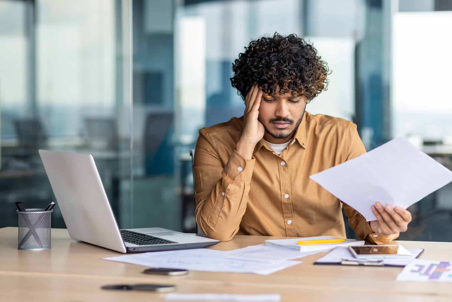 upset financier looking at sales and revenue chart of company, businessman inside office disappointed with sales results and achievements, hispanic man sitting at table with documents and laptop